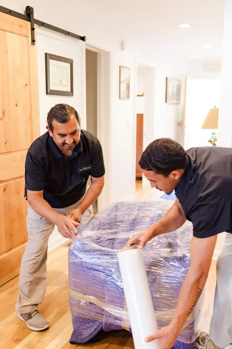 Two Gasperson movers wrapping a large item in a residential home